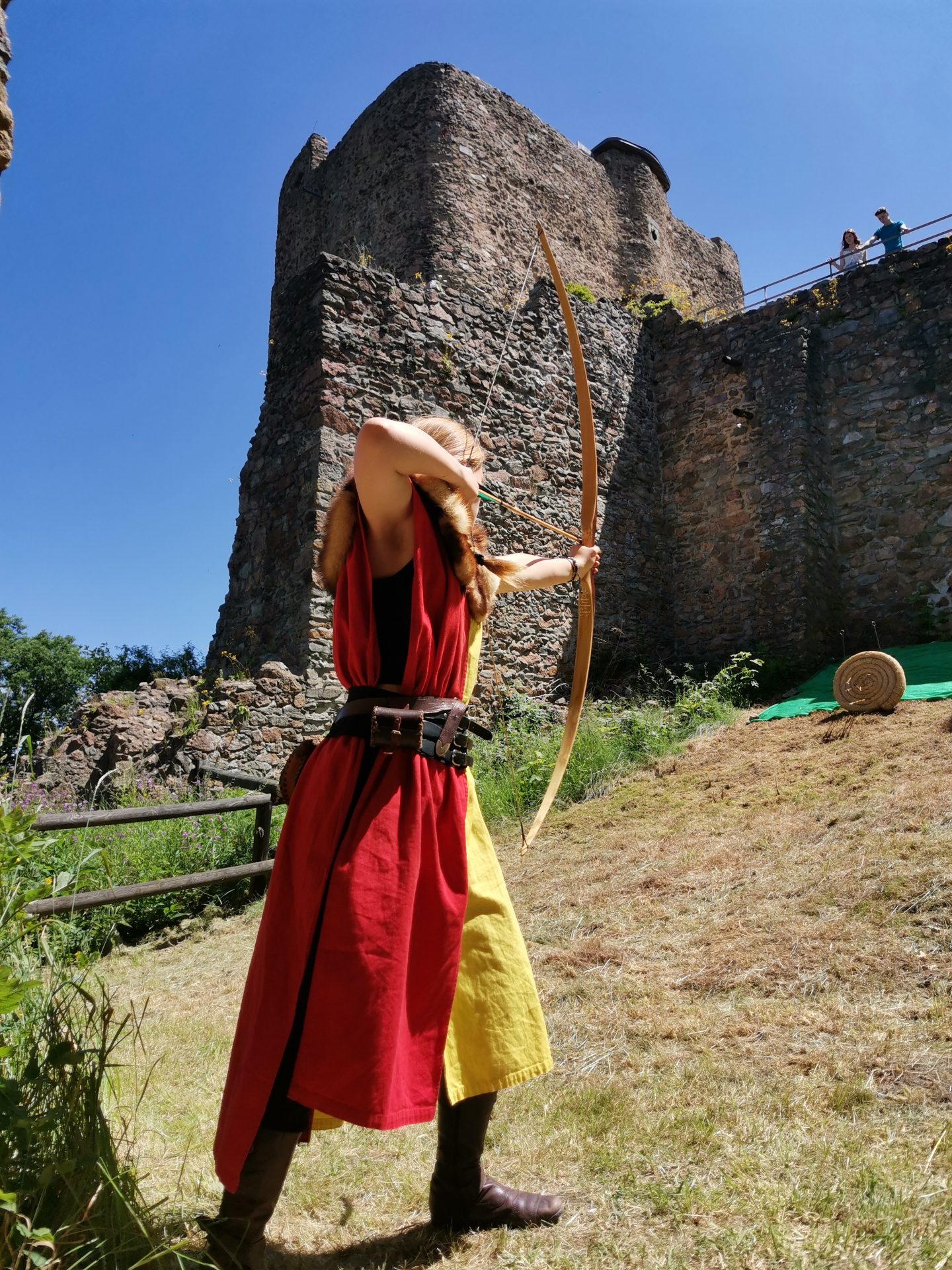 Bogenschützin in rotem mittelalterlichem Gewand zielt mit einem Langbogen vor dem Burgturm