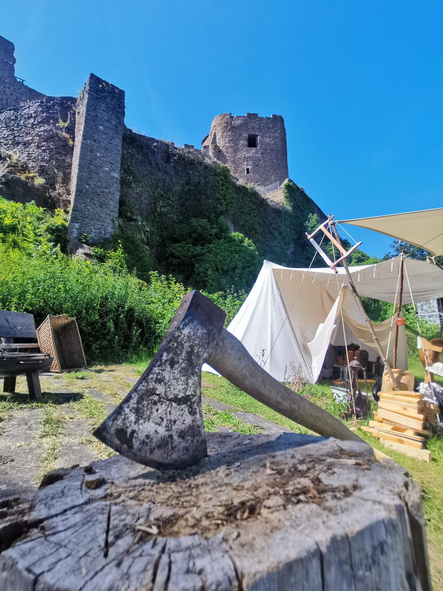 Mittelalterliche Axt steckt in einem Holzklotz, dahinter Lagerzelte und der Bergfried