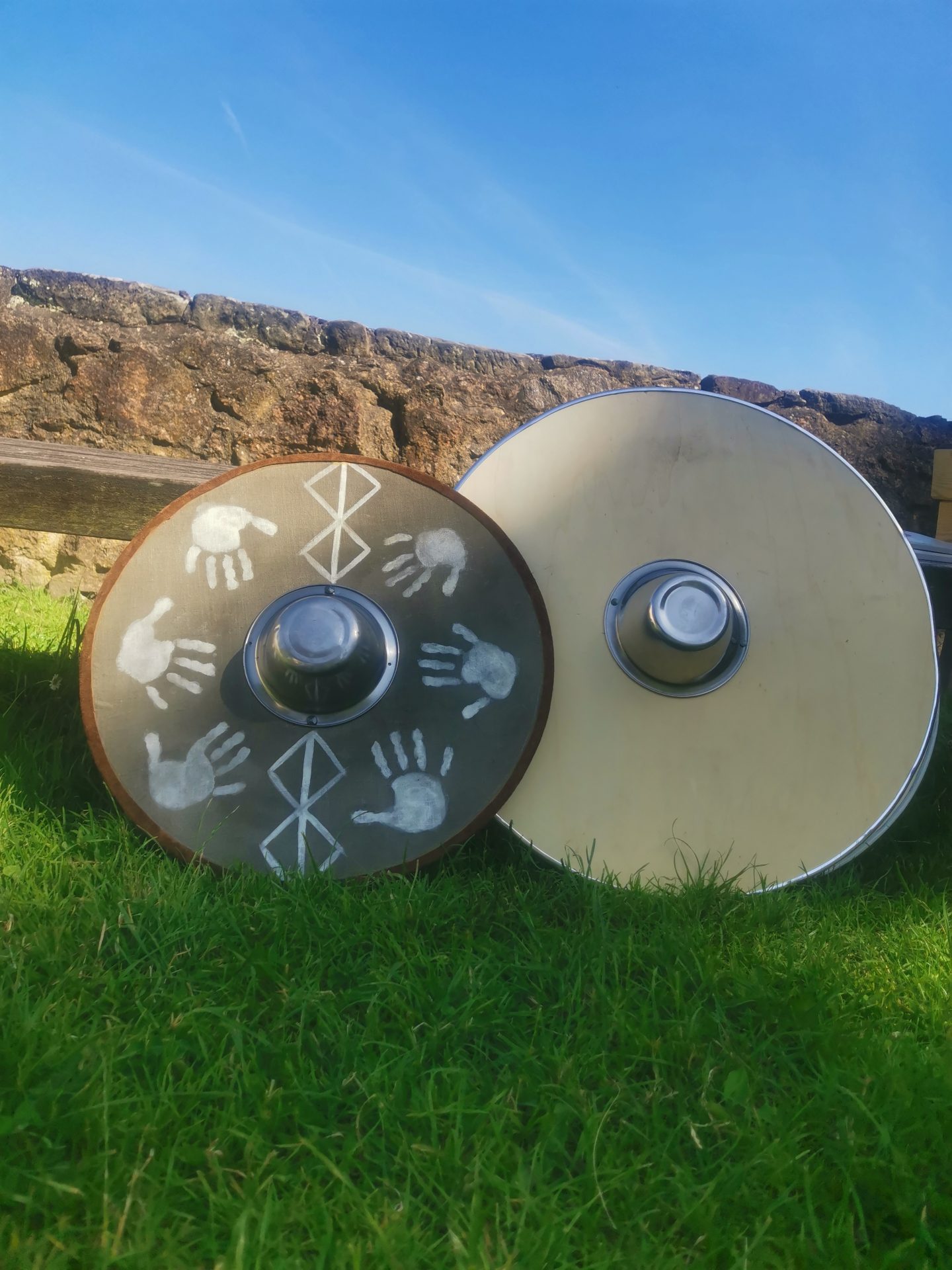 Zwei mittelalterliche Rundschilde mit Handabdrücken und Runen lehnen im Gras an einer Burgmauer