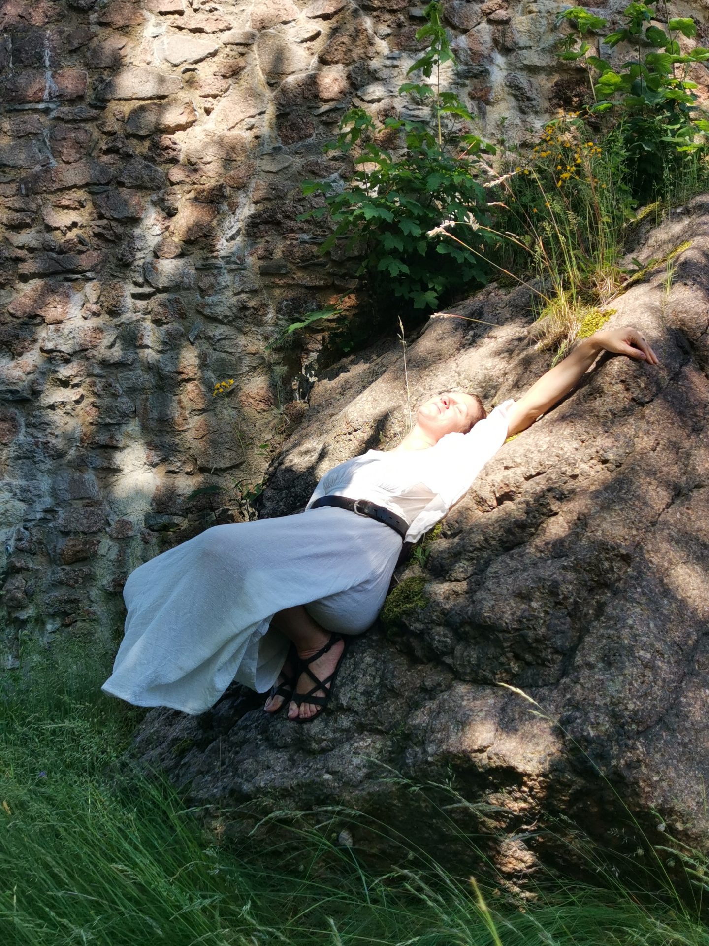 Darstellerin in weißem Gewand liegt entspannt auf einem Felsen an der alten Burgmauer